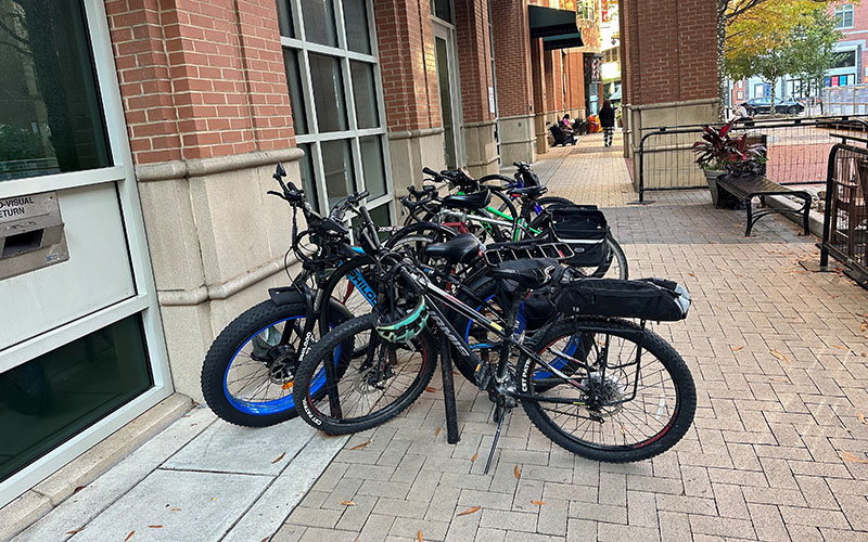 Several bicycles are parked and locked to a bike rack on a brick sidewalk next to a building with large windows and red brick walls.