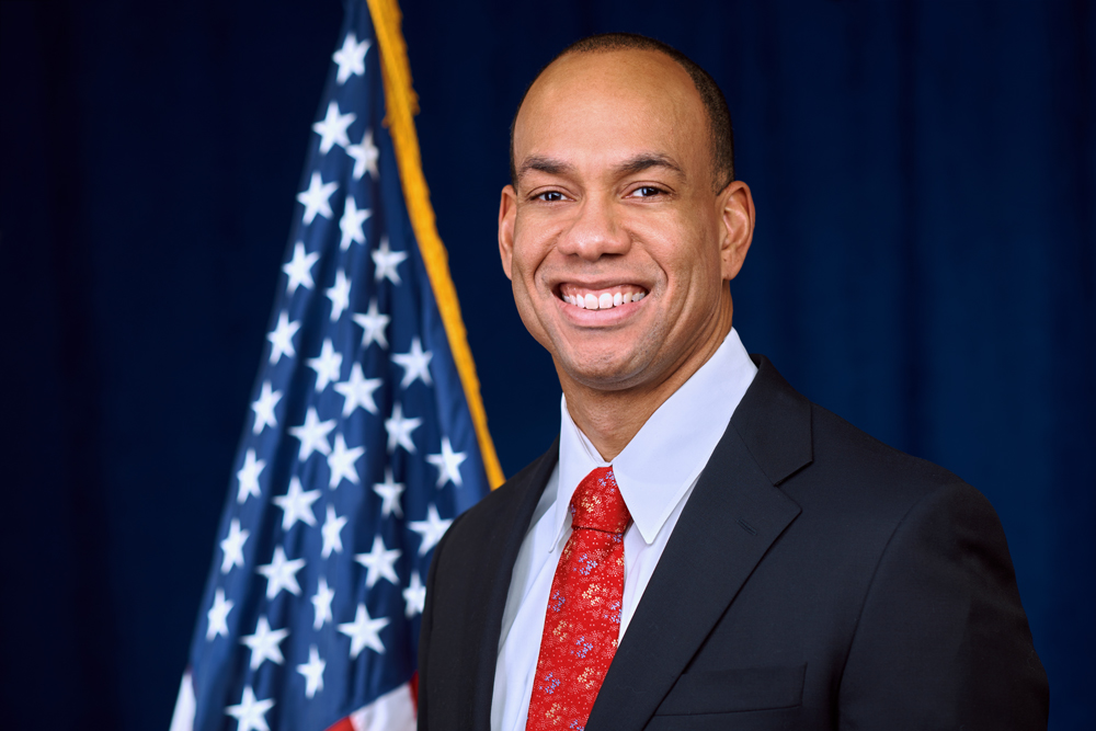 A man in a suit and red tie smiles in front of a U.S. flag and a blue background.