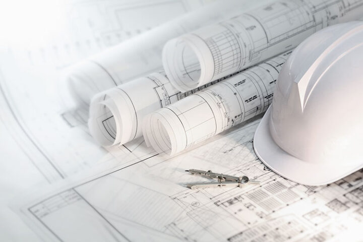 A white safety helmet, rolled architectural blueprints, and a compass rest on detailed construction plans.