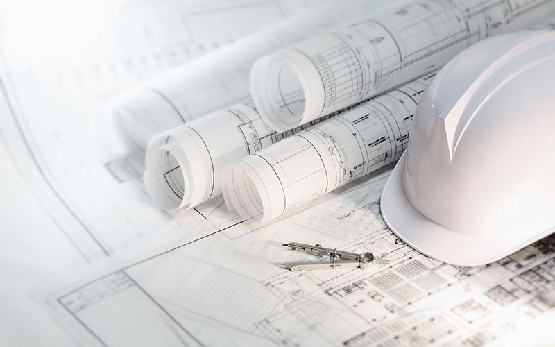 A white safety helmet, rolled architectural blueprints, and a compass rest on detailed construction plans.