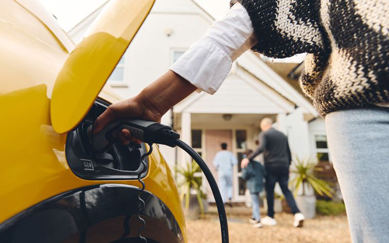 A person plugs a charging cable into a yellow electric car parked outside a house, with a family walking toward the front door in the background.