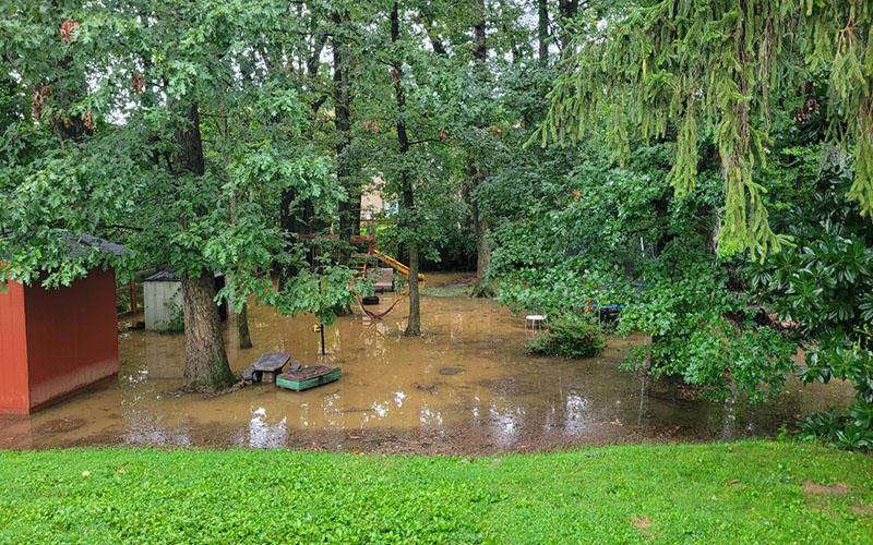 A backyard with grass, trees, a shed, and playground equipment is flooded with muddy water after heavy rain.