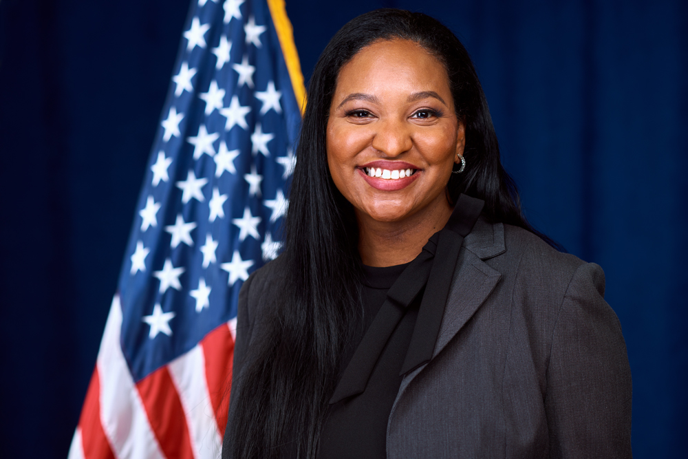 A woman in a dark suit smiles in front of an American flag and a blue backdrop.