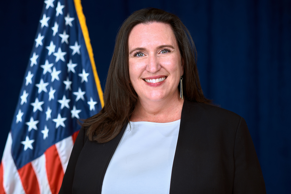 A woman in professional attire smiles in front of a U.S. flag and a dark blue background.