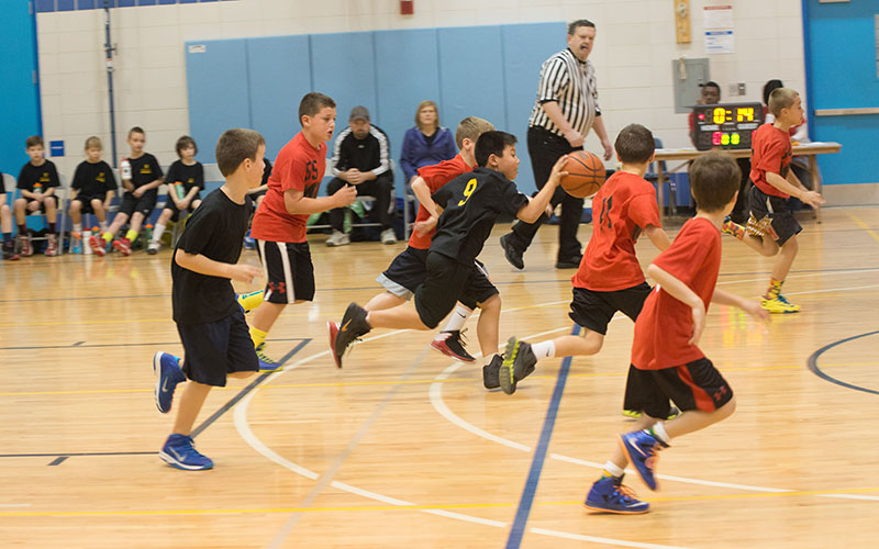 Children play a basketball game in a gymnasium, with one child dribbling the ball while others run beside him. A referee and spectators are visible in the background.