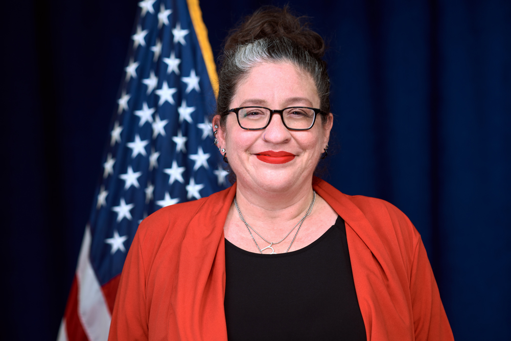 A woman with glasses and red lipstick stands in front of a U.S. flag, wearing a red jacket over a black top, against a dark blue background.