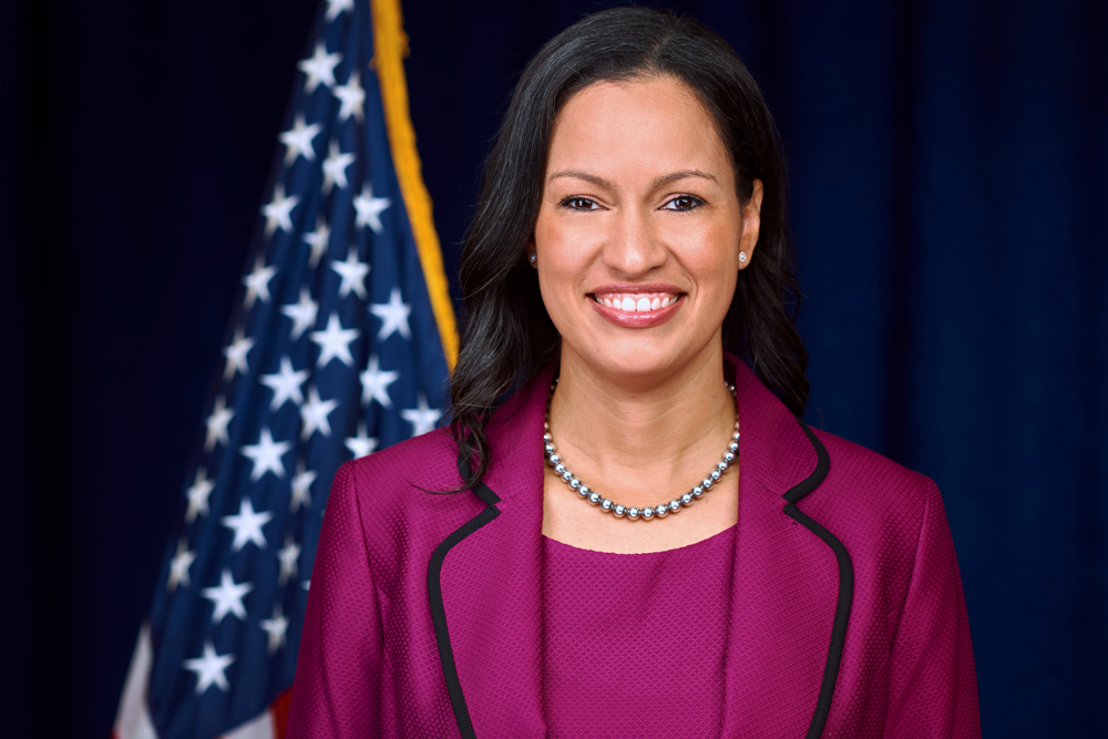 A woman in a magenta suit and pearl necklace stands smiling in front of a dark blue backdrop with the United States flag visible behind her.