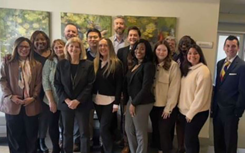 A group of fourteen people, dressed in business attire, pose together indoors in front of abstract artwork.