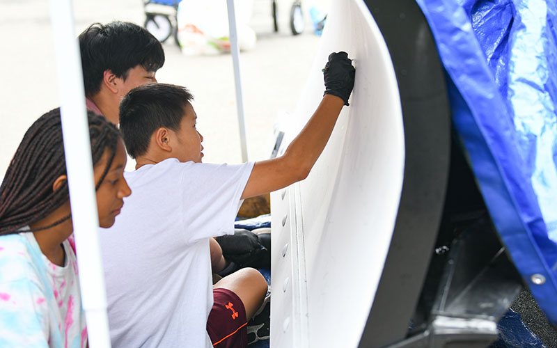 Three young people work together on a large white object, with one wearing gloves and appearing to sand or smooth its surface.