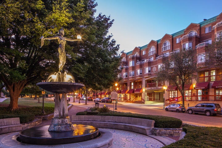 A bronze statue stands atop a fountain in a small plaza, with parked cars and a multi-story brick building with storefronts in the background at dusk.