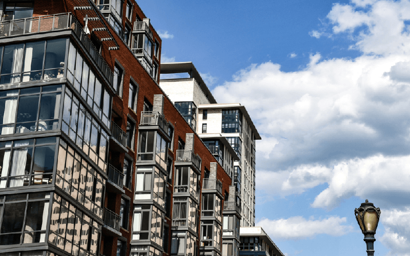 Multi-story modern apartment building with glass windows and balconies, adjacent to a streetlamp, set against a blue sky with scattered clouds.