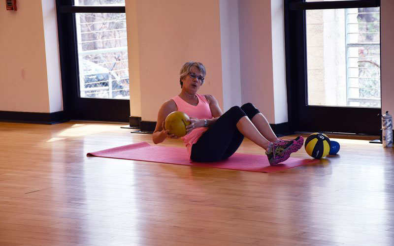 A woman sits on a yoga mat in a gym, holding a medicine ball and twisting her torso, with additional medicine balls nearby.