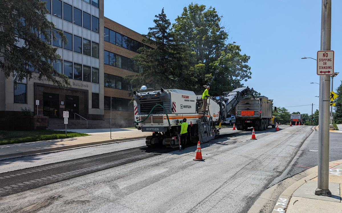 Road construction crew operates heavy machinery to resurface an urban street, with orange cones marking the work zone near an office building on a sunny day.
