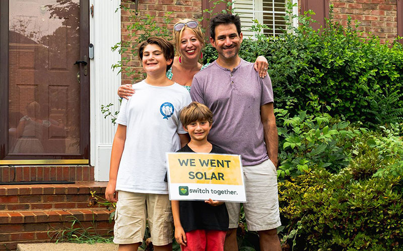 A family of four stands outside their house, smiling, with one child holding a sign that reads, "WE WENT SOLAR switch together.
