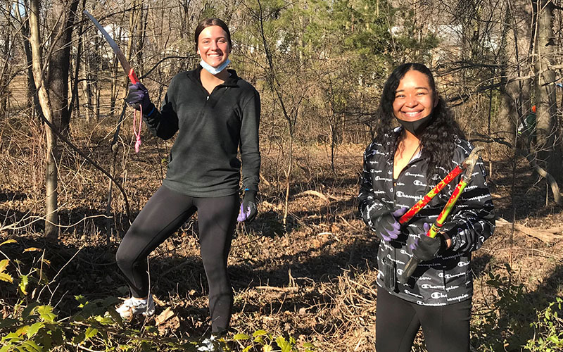 Two women stand outdoors in a wooded area, smiling and holding gardening tools while dressed in casual clothing and gloves.