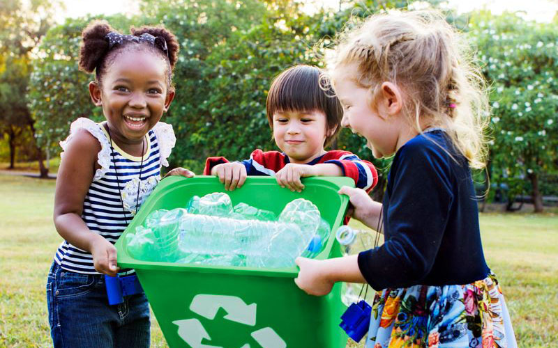 Three children hold a green recycling bin filled with plastic bottles outdoors, smiling and participating in a recycling activity.