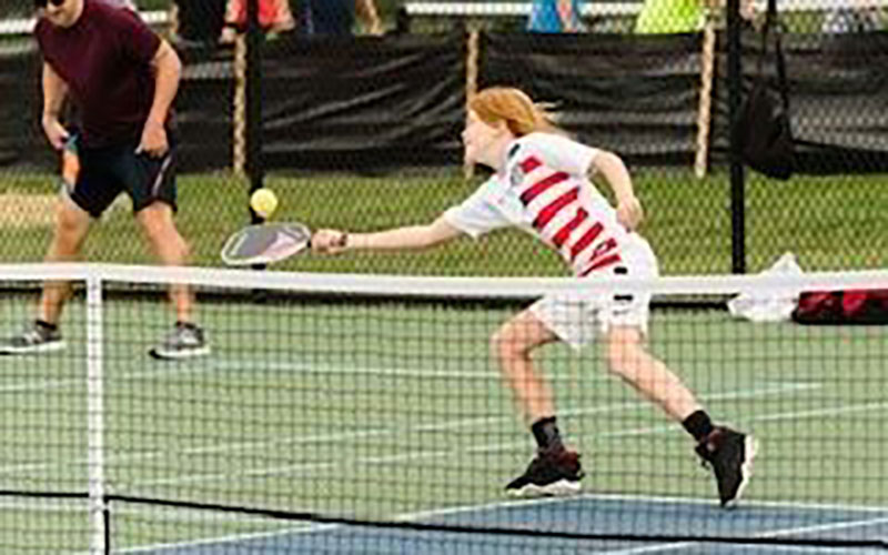 A person lunges forward on an outdoor court to hit a pickleball with a paddle, while another player stands in the background.
