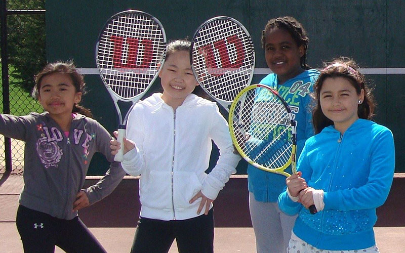 Four girls stand on a tennis court holding tennis rackets, smiling at the camera. A fence and green wall are visible in the background.