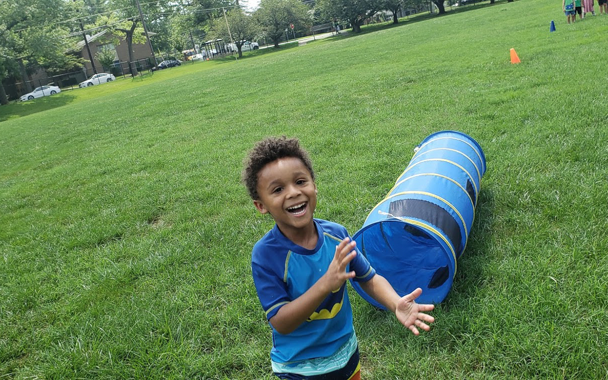 A young boy smiles and claps near a blue play tunnel on a grassy field, with trees and cones visible in the background.