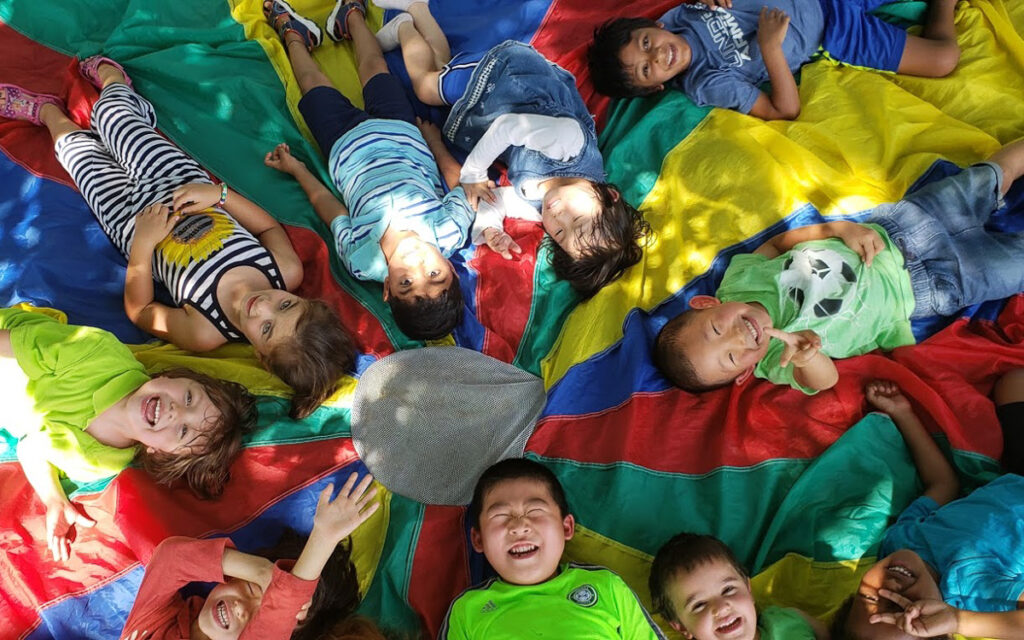 A group of children lie in a circle on a colorful parachute, smiling and looking up at the camera.