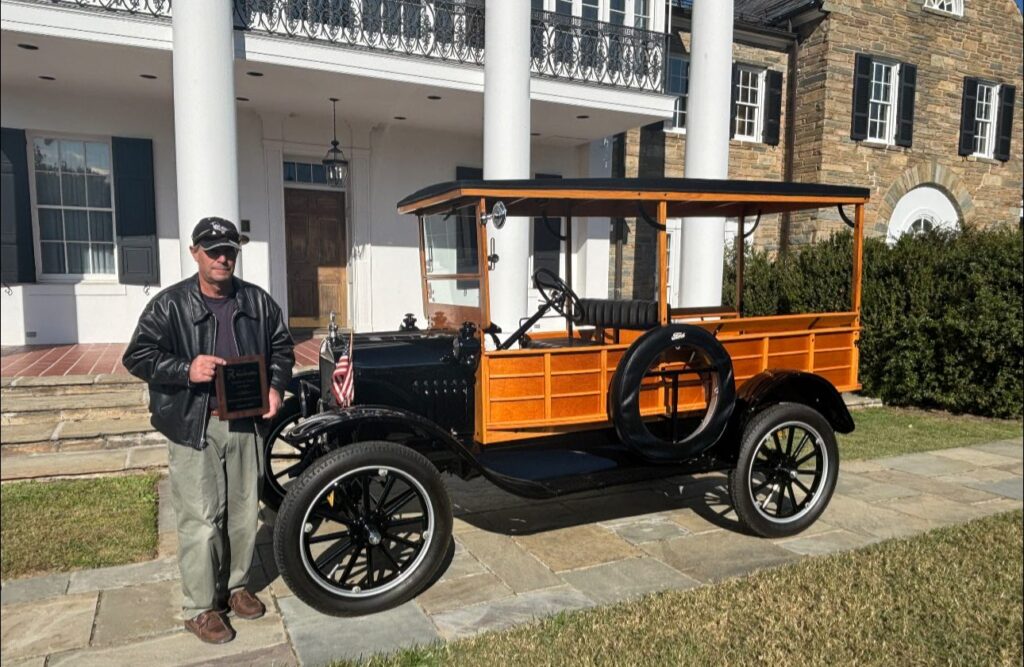 A person in a leather jacket and cap stands next to a vintage wooden truck, holding a plaque, in front of a large stone and brick house with white pillars.