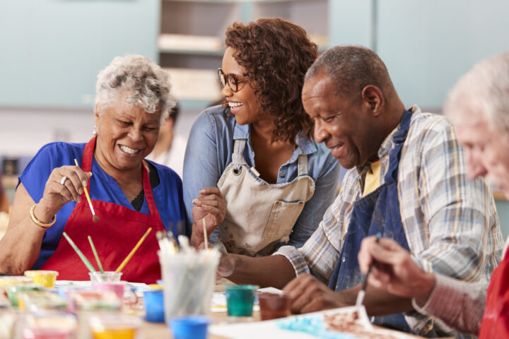 Four adults sit at a table painting and smiling, with art supplies spread out in front of them in a brightly lit room.