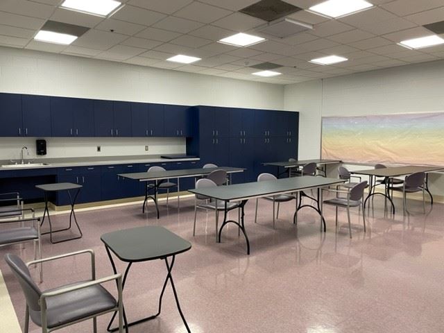 Classroom with several tables and chairs arranged in small groups, blue cabinets along the wall, and a pastel mural on the right. The room is brightly lit with overhead lights.