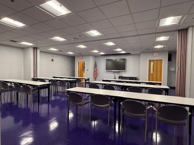 A classroom or meeting room with rows of tables and chairs, a large wall-mounted screen, an American flag, and fluorescent ceiling lights.