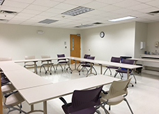 A conference room with empty tables arranged in a U-shape, several chairs, a wall clock, a closed door, and a sink area in the corner.