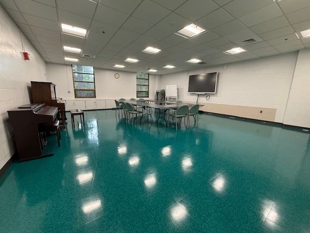A classroom with turquoise flooring, a piano, tables and chairs in the center, a large wall-mounted monitor, windows, and fluorescent ceiling lights.