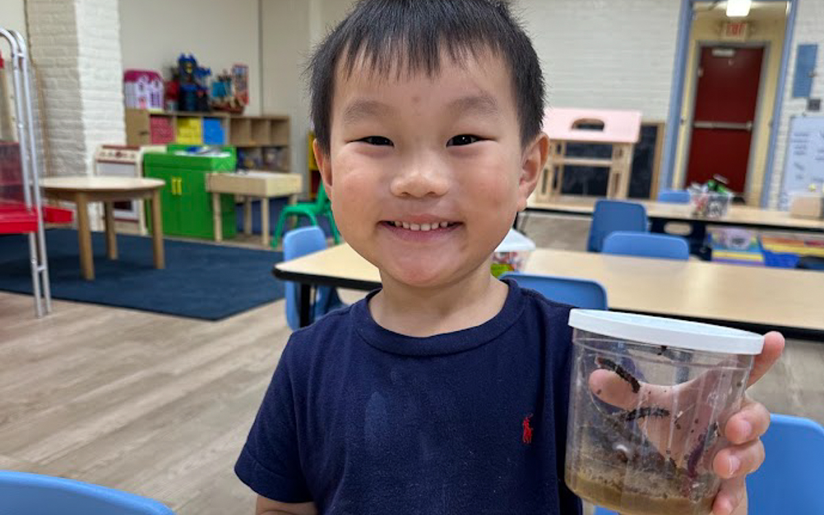 A young child smiles while holding a clear container with soil and worms inside, standing in a classroom with tables and chairs.