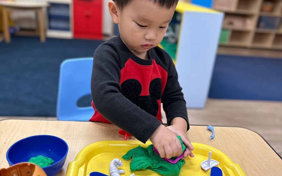 A young child presses green playdough with a cutter on a yellow tray, seated at a table in a classroom setting.