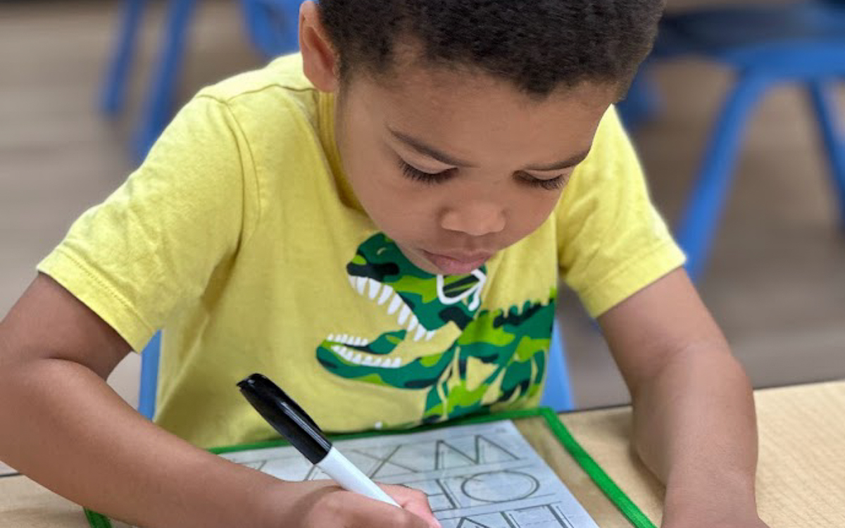 A young child in a yellow dinosaur shirt practices writing letters with a marker on a laminated worksheet at a classroom table.