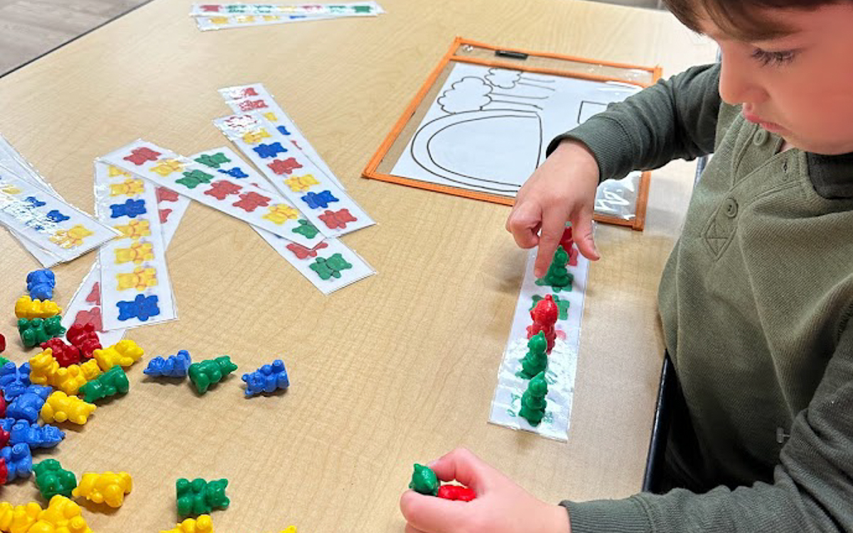 A child arranges colorful plastic bear counters on a pattern strip at a table covered with similar strips and bears.