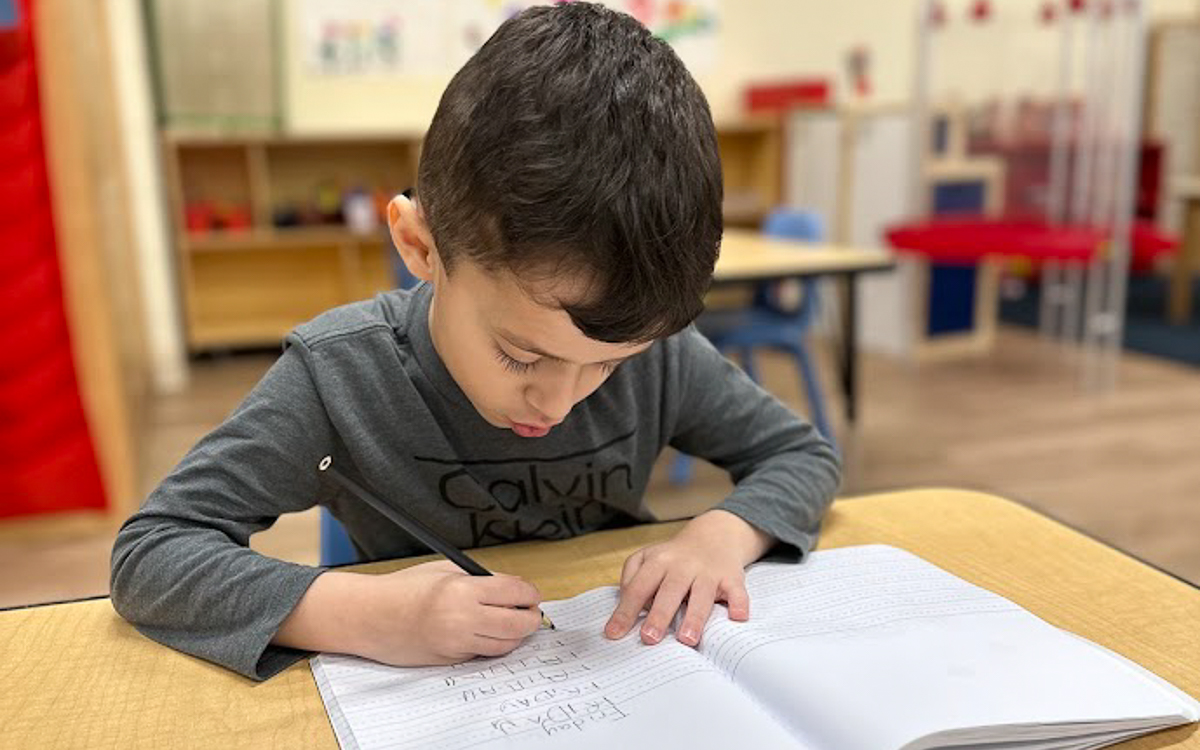 A young boy sits at a desk in a classroom, writing in a notebook with a pencil.