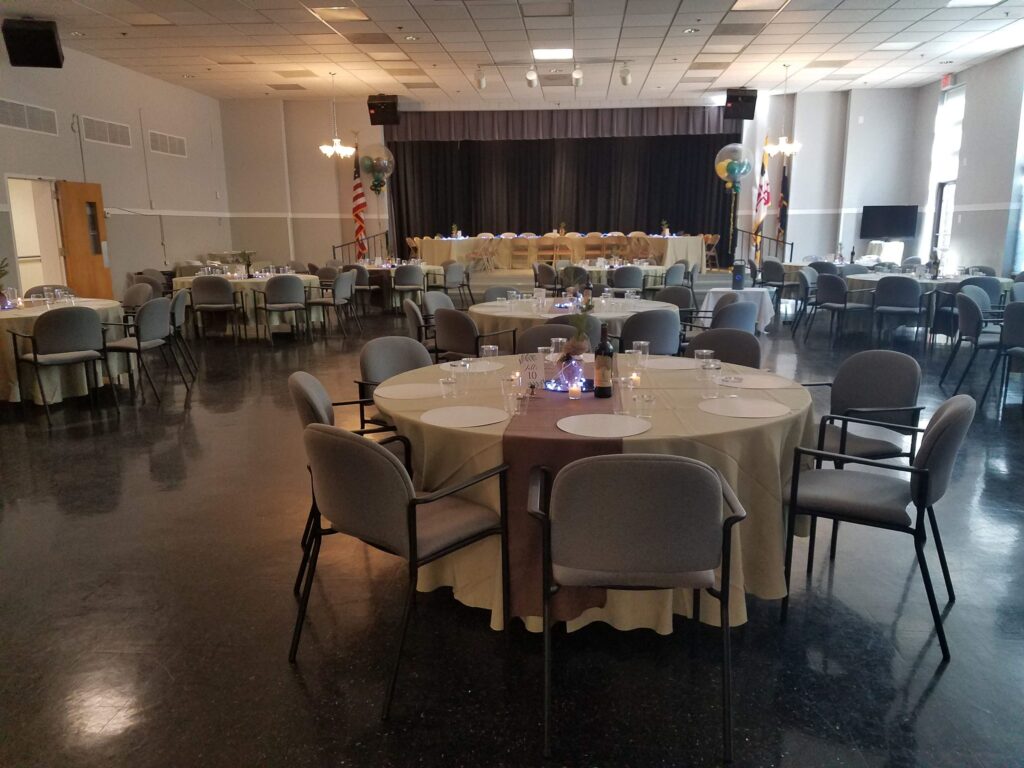 A banquet hall with round tables covered in beige tablecloths, set for an event; a stage with chairs and decorations is visible at the back of the room.
