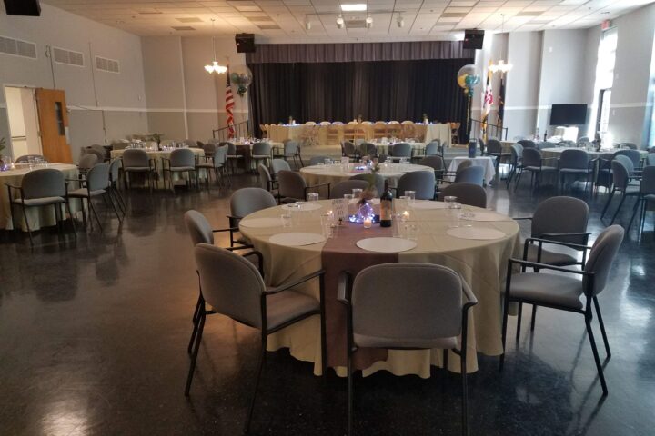A banquet hall with round tables covered in beige tablecloths, set for an event; a stage with chairs and decorations is visible at the back of the room.
