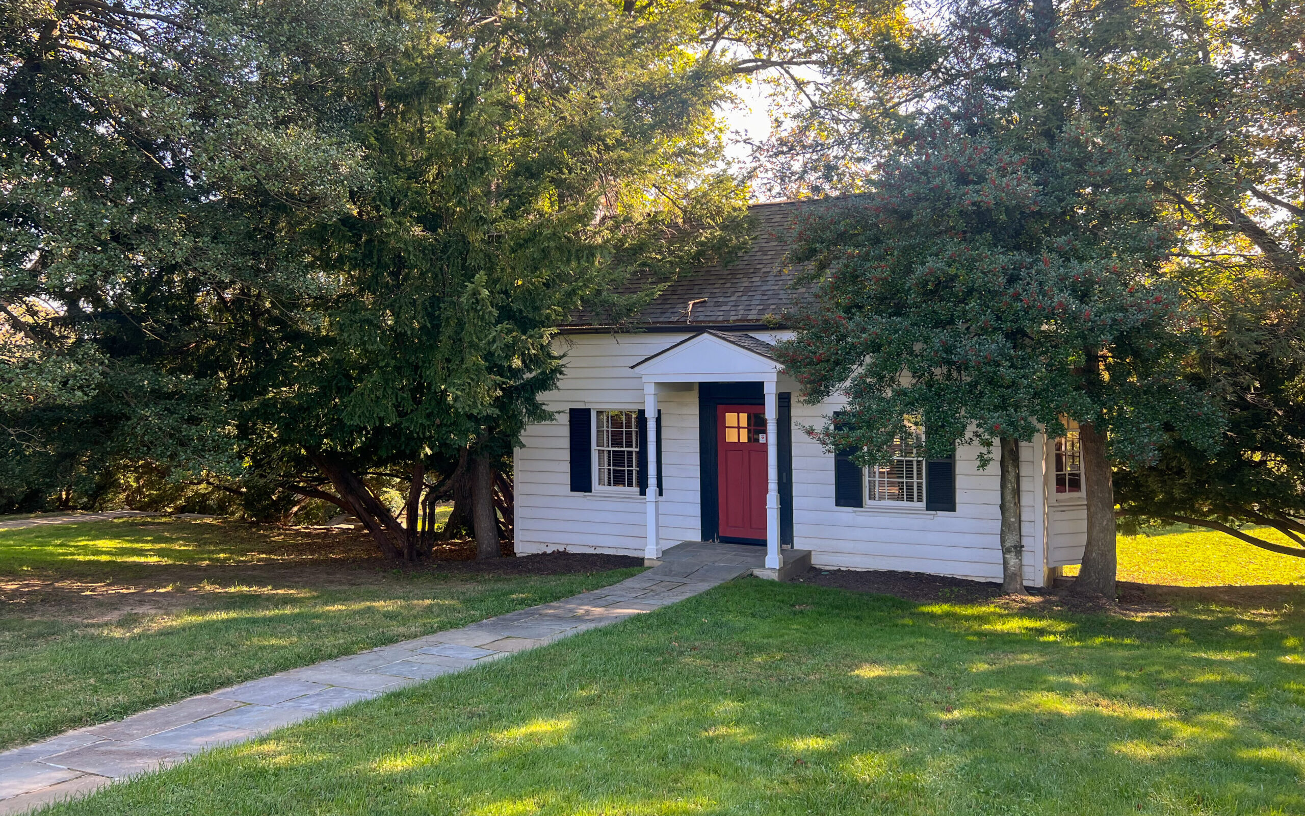 Small white house with a red front door, blue shutters, and a stone walkway, surrounded by large trees and green grass on a sunny day.