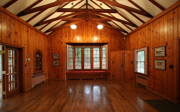 Spacious room with polished wooden floors, wood-paneled walls, exposed ceiling beams, large windows with blinds, and framed pictures on the walls.