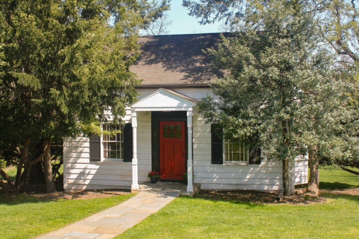 A small white house with black shutters and a red front door is surrounded by trees and a green lawn, with a stone walkway leading to the entrance.
