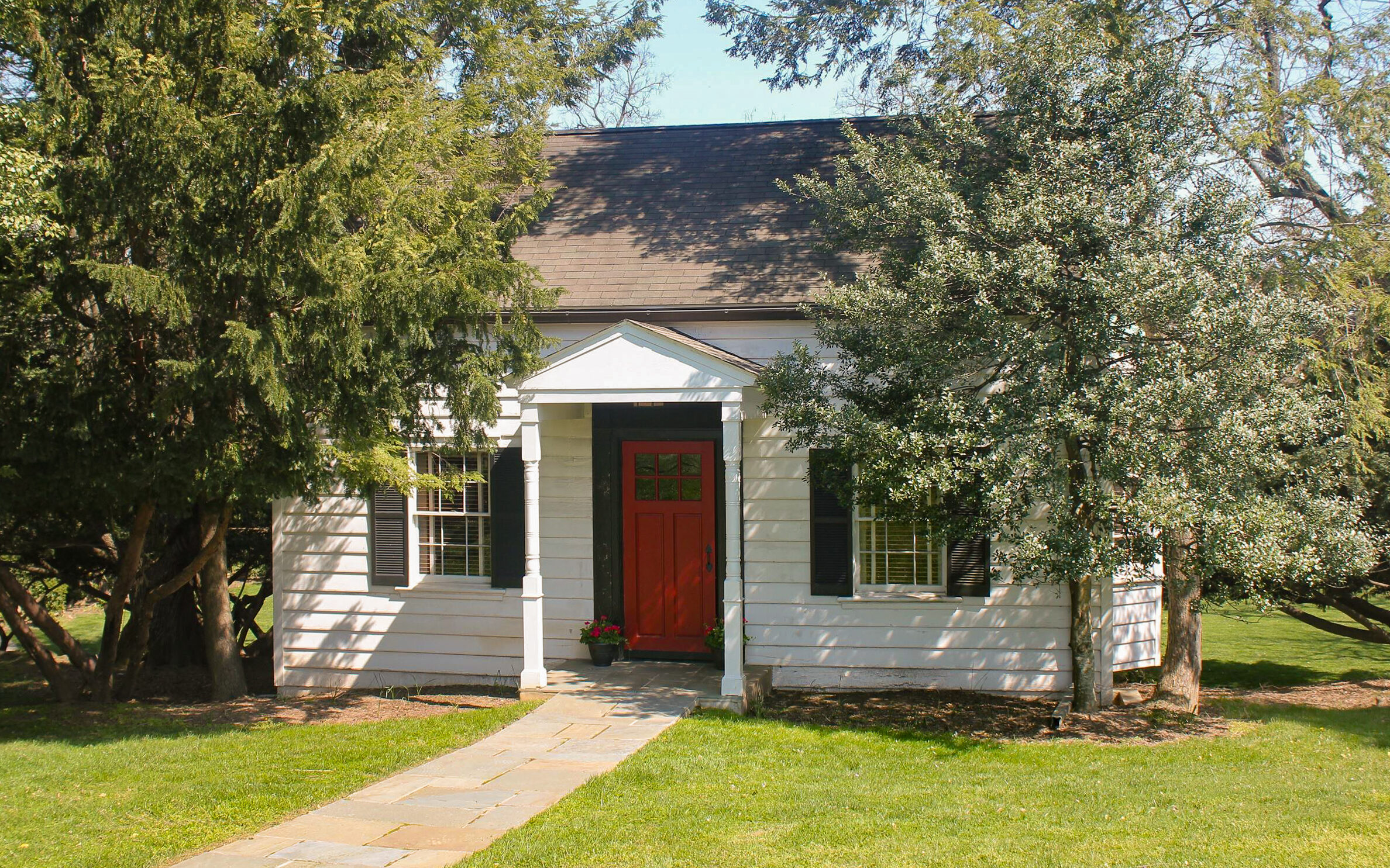 A small white house with black shutters and a red front door is surrounded by trees and a green lawn, with a stone walkway leading to the entrance.