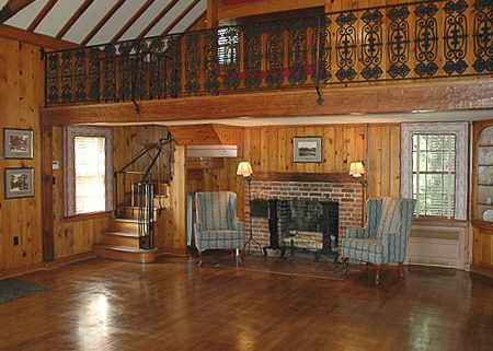 Wood-paneled living room with a brick fireplace, two striped armchairs, a spiral staircase, wooden floors, and a decorative iron railing on the loft.