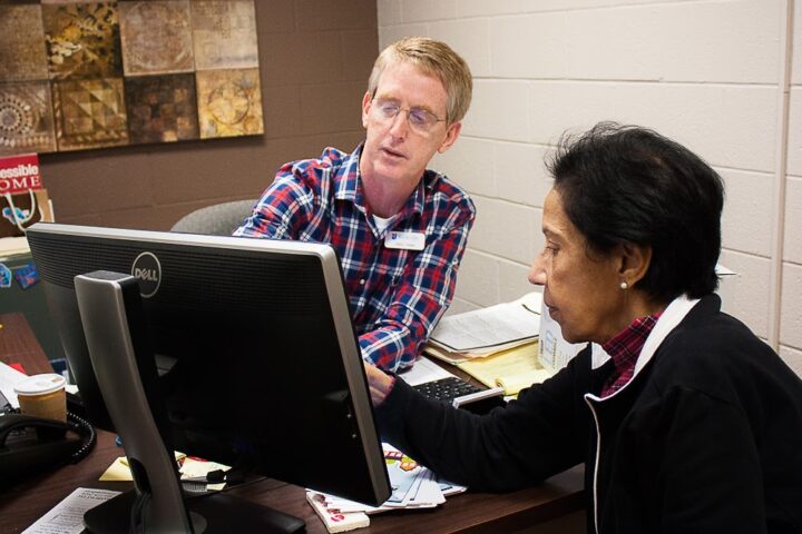 Two people sit at a desk looking at a computer monitor together; the man is pointing at the screen while the woman watches attentively.