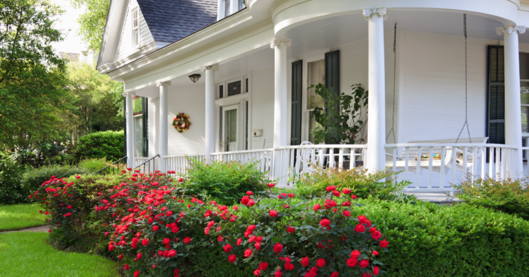 A white house with a wraparound porch, white columns, a porch swing, and red flowering bushes in a green, landscaped yard.