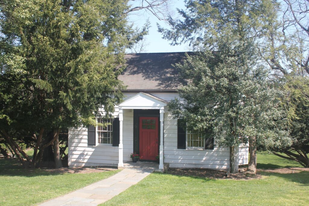 A small white house with black shutters and a red front door is surrounded by tall green trees and sits on a lawn with a stone pathway.
