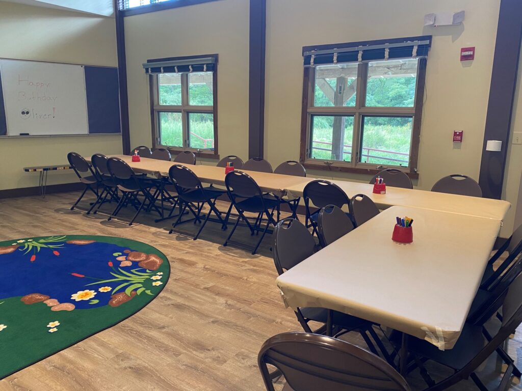 A classroom with two long tables and chairs, a rug with a flower design, and art supplies on the tables. Large windows let in natural light. A birthday message is written on the chalkboard.