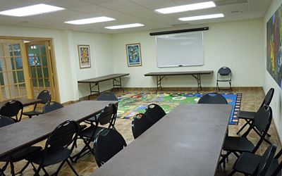 A classroom with folding tables and chairs, a whiteboard, artwork on the walls, and a colorful rug on the floor.