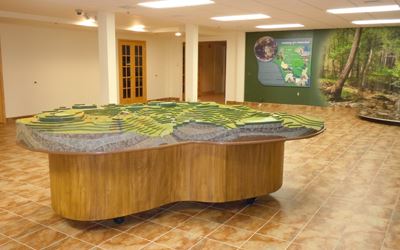 A three-dimensional topographic model of a green landscape is displayed on a wooden table in a tiled, well-lit room with informational posters on the walls.