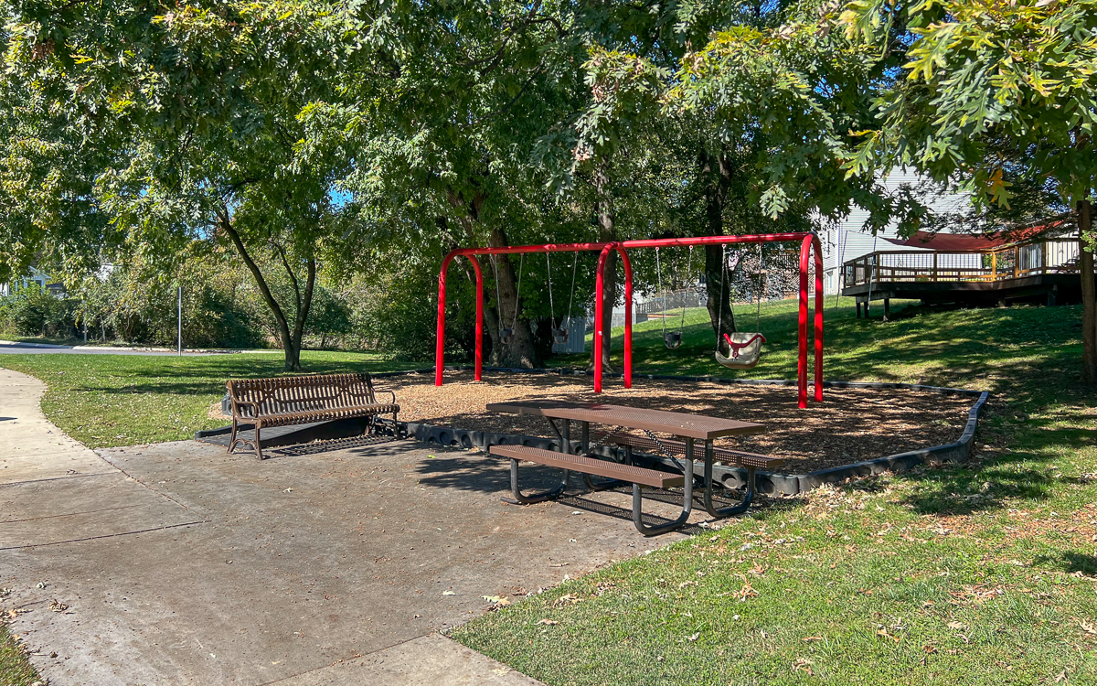 A playground with two red swings, a wooden bench, and a picnic table surrounded by green grass and trees on a sunny day.