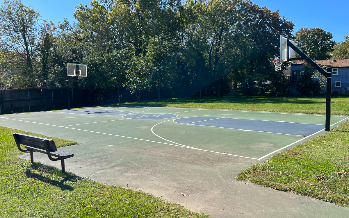 Outdoor basketball court with two hoops, surrounded by grass and trees, featuring a bench in the foreground. The sky is clear and sunny.